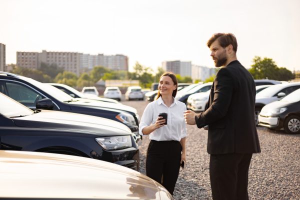 A male car dealer discusses options with a female customer at a car dealership.
